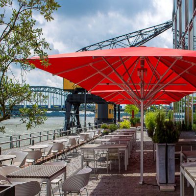 Rote Sonnenschirme für die Gastronomie mit Beleuchtung und Heizstrahlern, auf einer Terrasse am Wasser mit Blick auf eine Brücke und einen Kran.