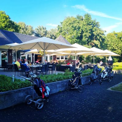 Eine Terrasse eines Golfclubs mit mehreren cremefarbenen Sonnenschirmen, umgeben von Grünflächen. Vor der Terrasse stehen Golftrolleys, während Gäste an Tischen entspannen.